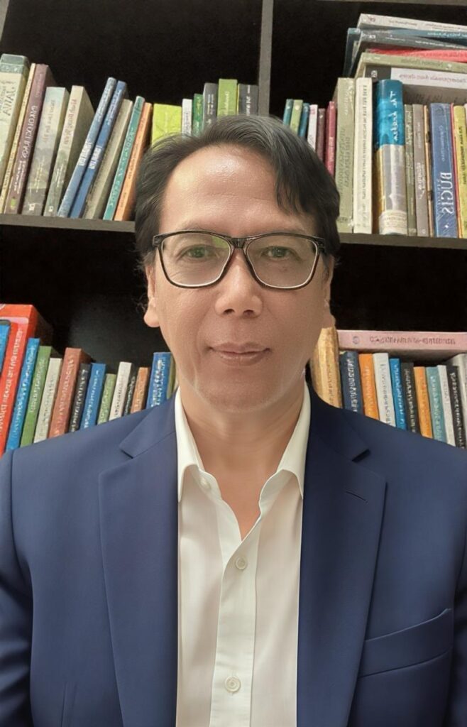 Sejarawa dr sri margana catrawarta Man in a navy blazer and white shirt wearing glasses standing in front of a colorful bookshelf